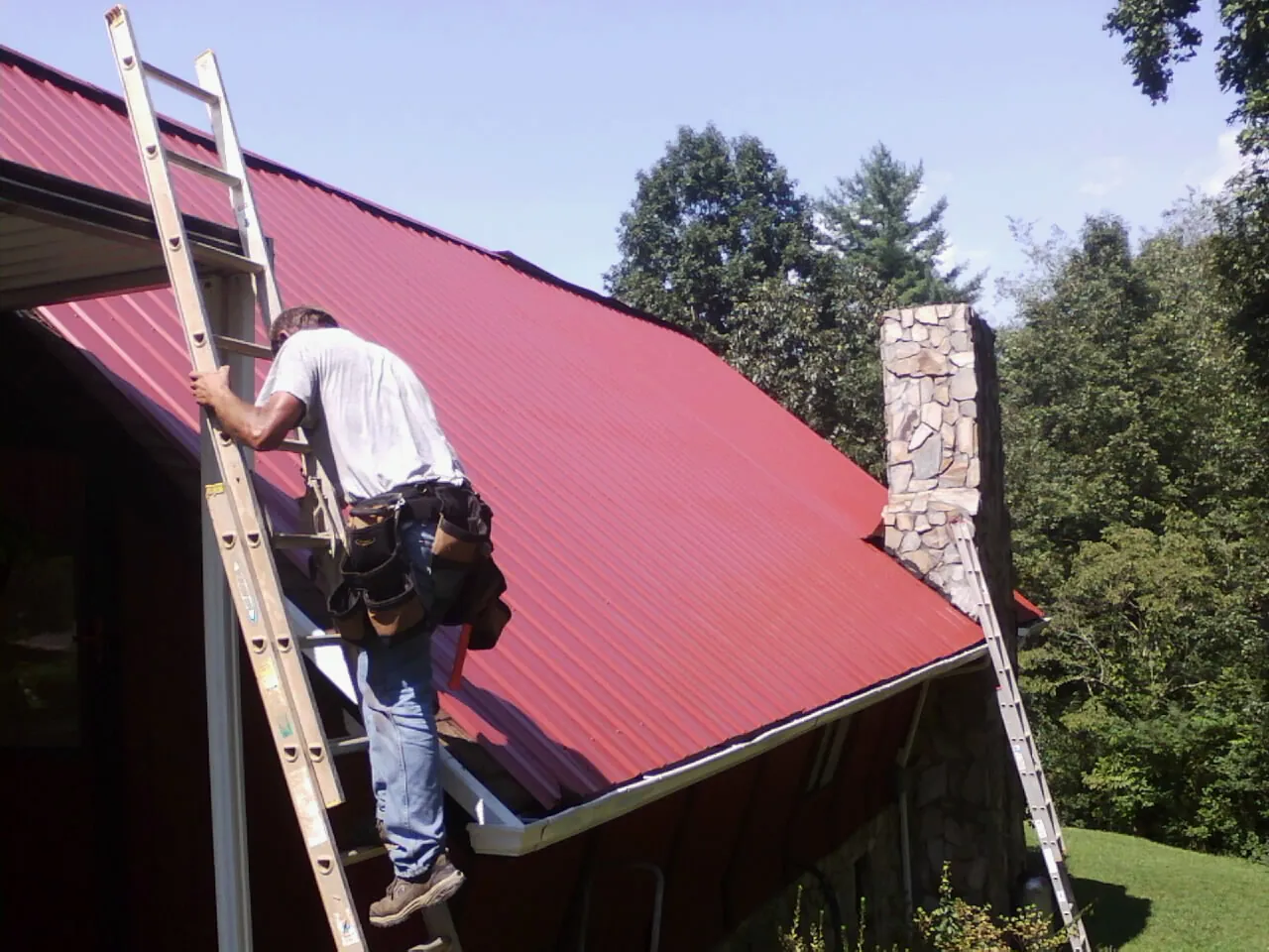 Roofer on a ladder beside a red metal roof and stone chimney — Soffit & Fascia Repair by Roofing Brothers in Rocky Top